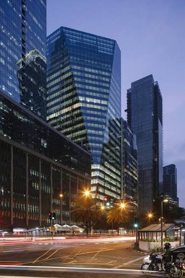Nighttime street scene with tall glass skyscrapers, illuminated windows, palm trees, and light trails from passing cars.
