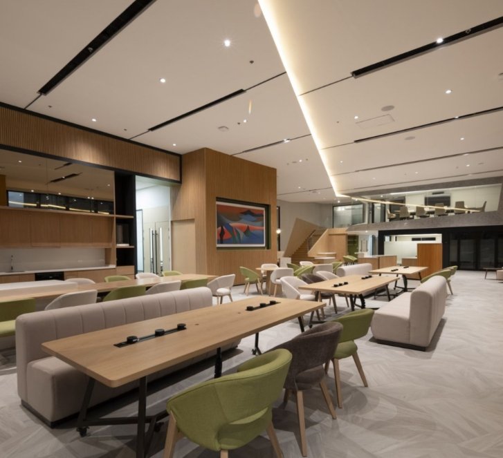 Open, modern coworking space with long wooden tables, lounge chairs, and artwork on wood-paneled walls in a bright setting.