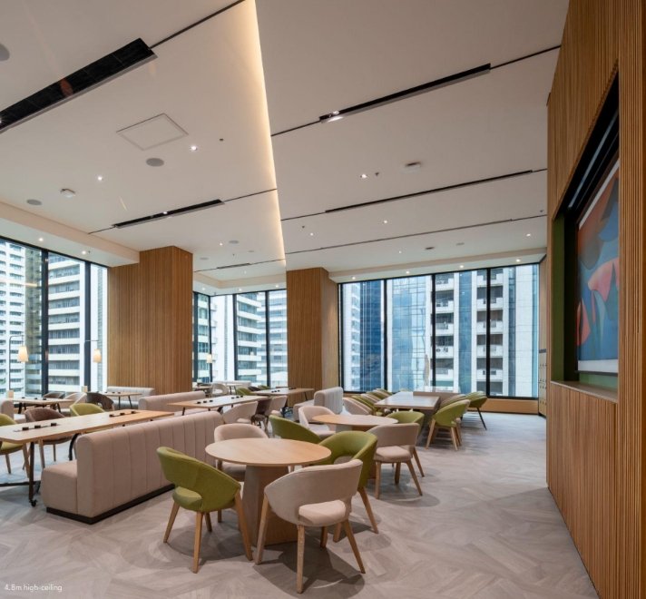 Modern office lounge with round wooden tables, green and beige chairs, and light wood dividers under a high, bright ceiling, with floor-to-ceiling windows showing city buildings.