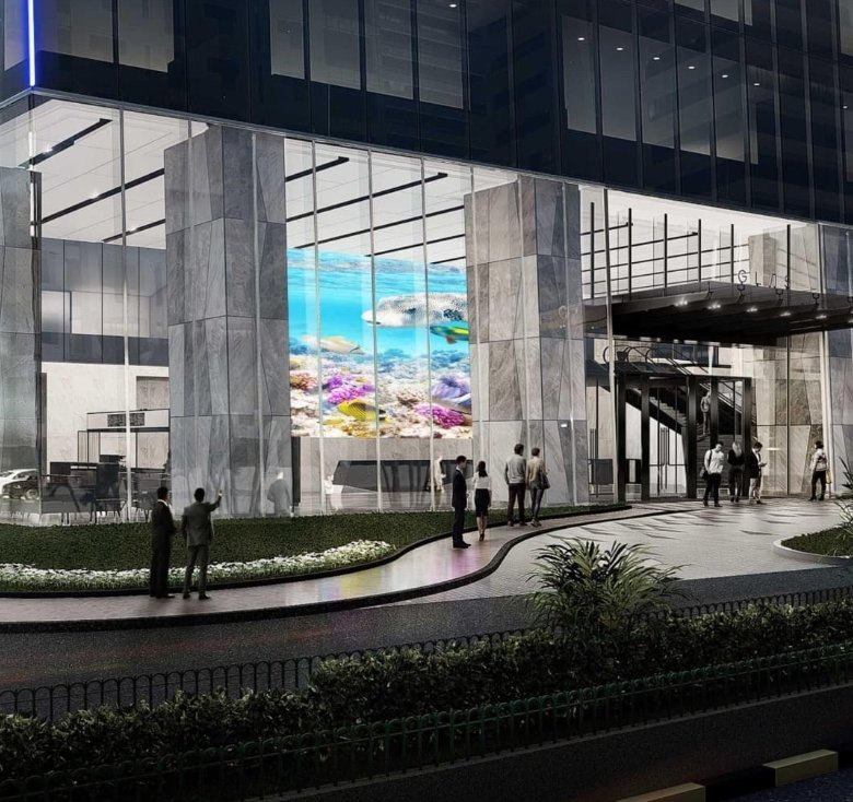 Nighttime view of a glass modern building with a covered drop-off area, pedestrians on the sidewalk, and cars along the curb; a bright aquarium-style screen is visible inside. The word 'Drop Off' appears at left.