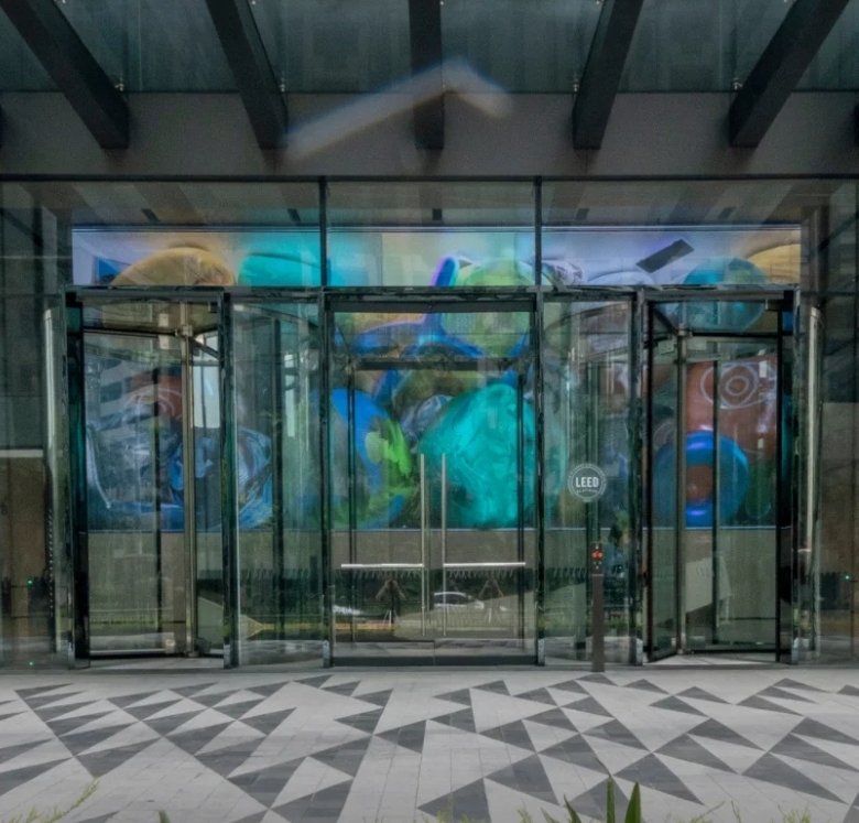 Modern glass building entrance with revolving doors and a geometric tiled plaza; two pedestrians on the right; 'Drop Off' label on the left.