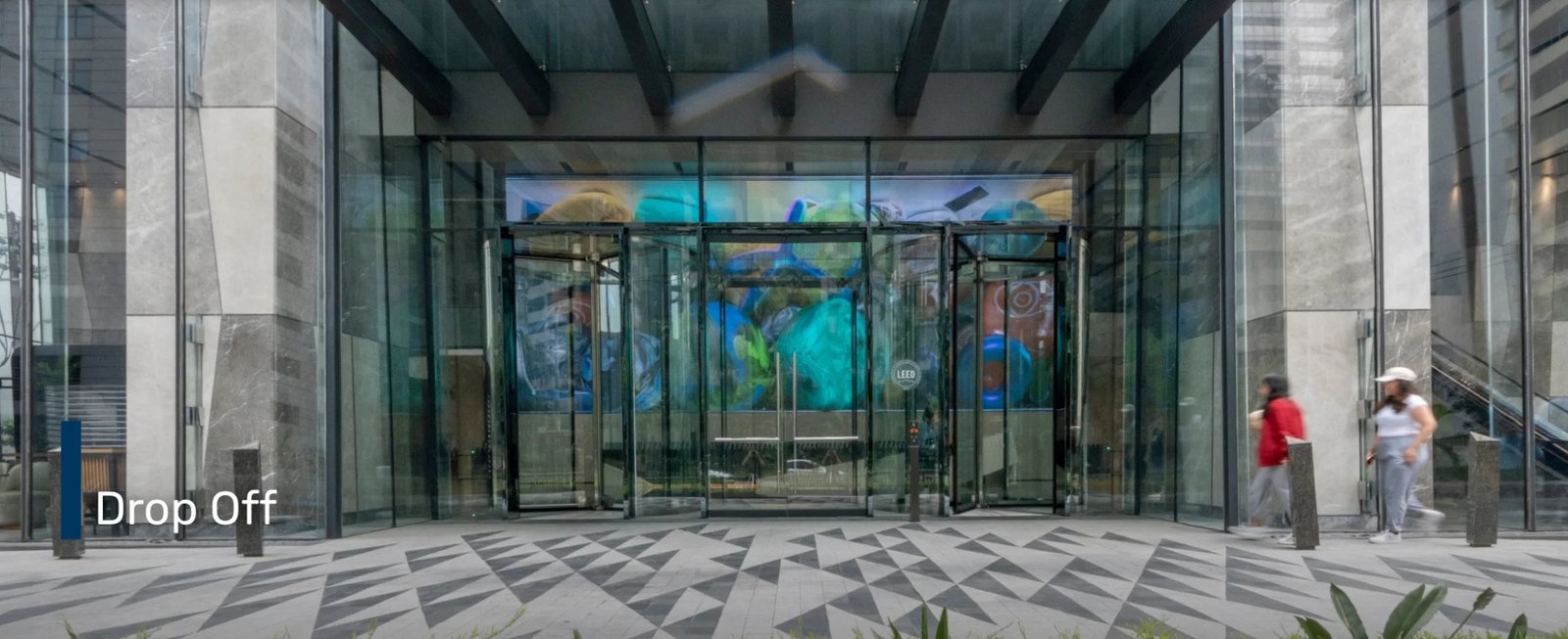 Screenshot_18-6-2026_172111_glas.ph Modern glass building entrance with revolving doors and a geometric tiled plaza; two pedestrians on the right; 'Drop Off' label on the left.