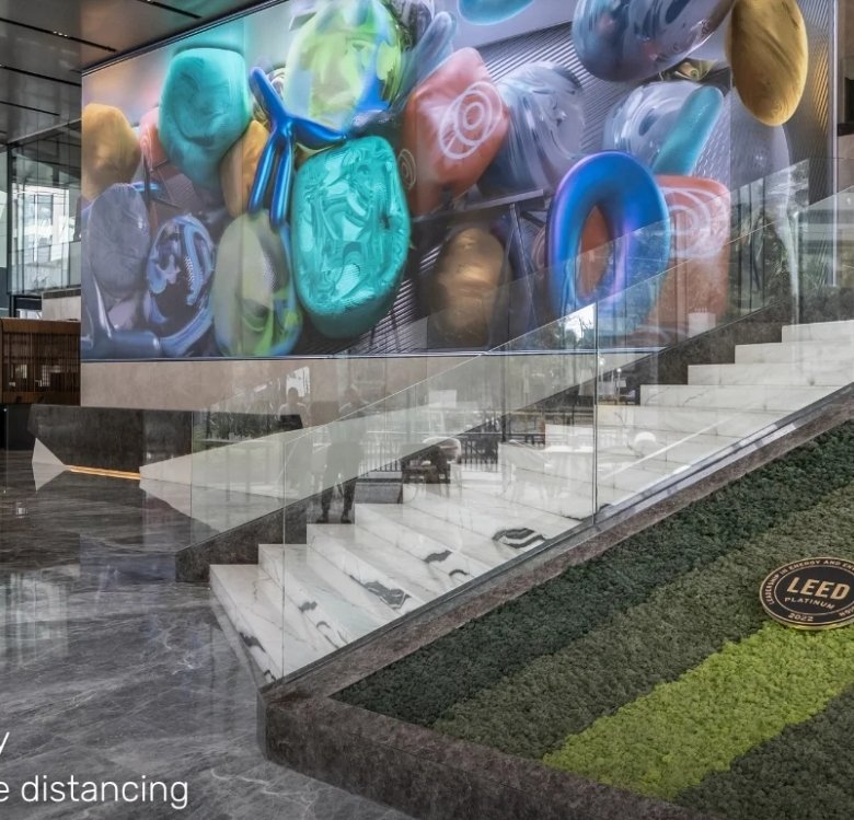 Modern hotel or office lobby with glass walls, a large colorful mural, and a green living wall beside an escalator on the right; Starbucks counter visible in the background.