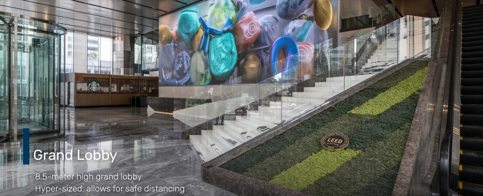 Screenshot_18-6-2026_172123_glas.ph Modern hotel or office lobby with glass walls, a large colorful mural, and a green living wall beside an escalator on the right; Starbucks counter visible in the background.