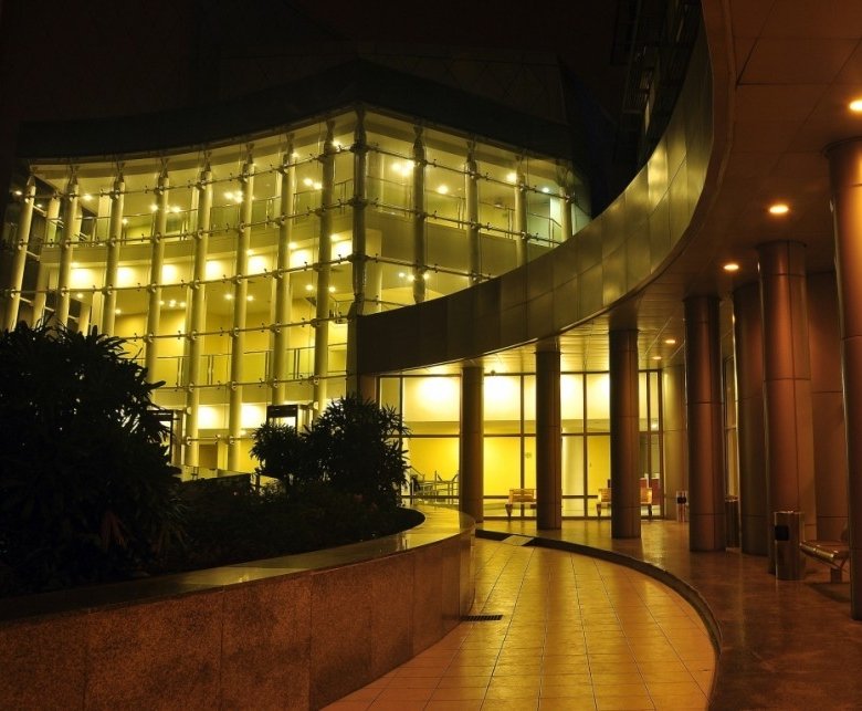 Night scene of a modern glass building with warm interior lights, a curved walkway, and tall columns.