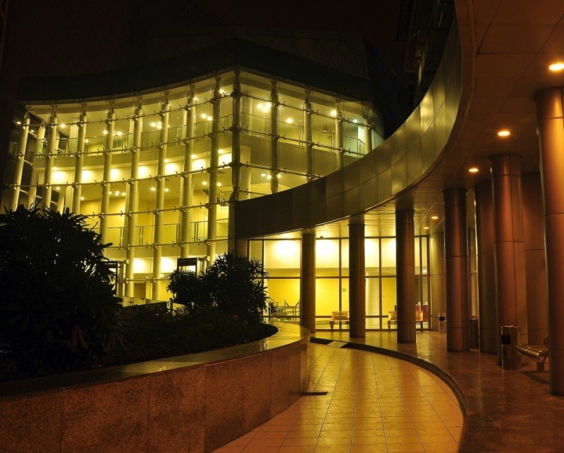 Night scene of a modern glass building with warm interior lights, a curved walkway, and tall columns.