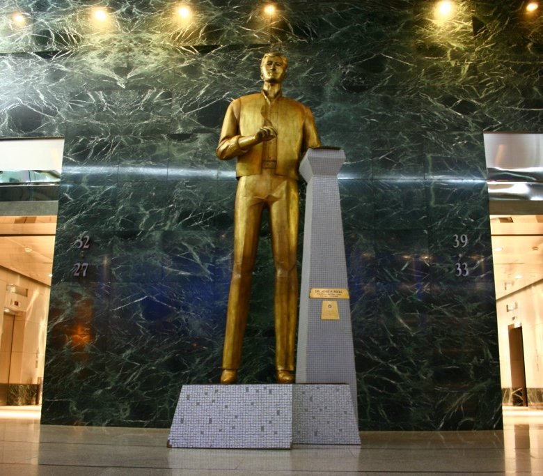Gold-colored statue of a man standing in a marble-walled lobby beside a tall obelisk pedestal and small mosaic base sculpture, with elevators visible on either side.