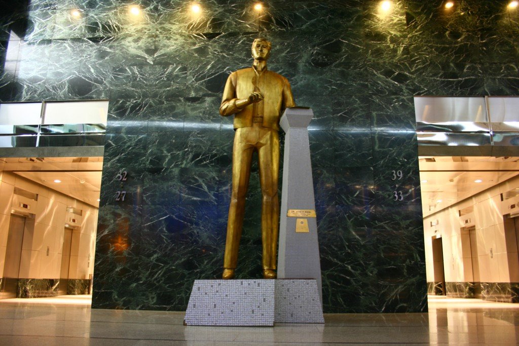 Gold-colored statue of a man standing in a marble-walled lobby beside a tall obelisk pedestal and small mosaic base sculpture, with elevators visible on either side.