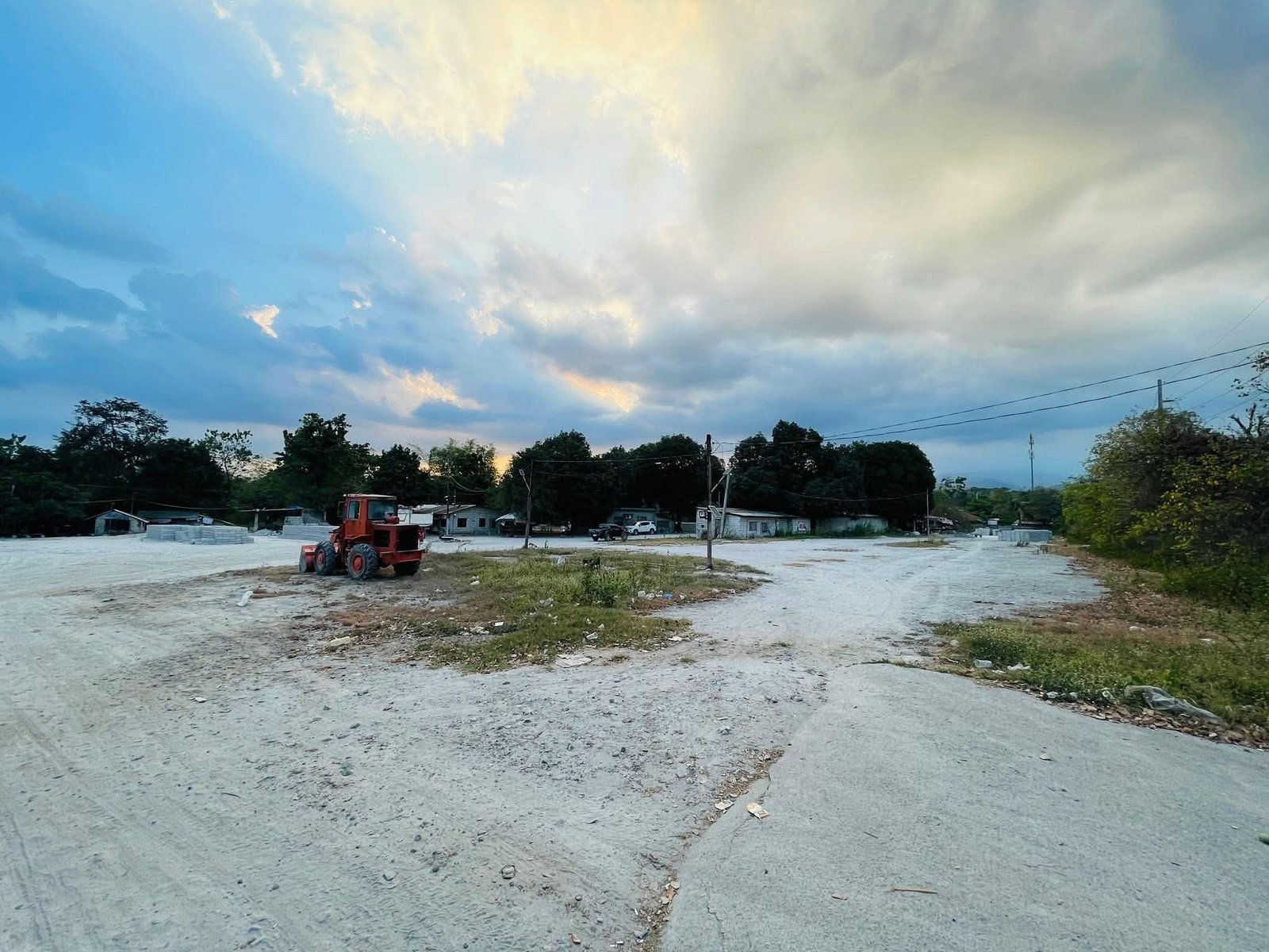Construction lot with a red heavy tractor on dusty ground and low-lying buildings along a tree line under a cloudy sky, power lines visible in the background.