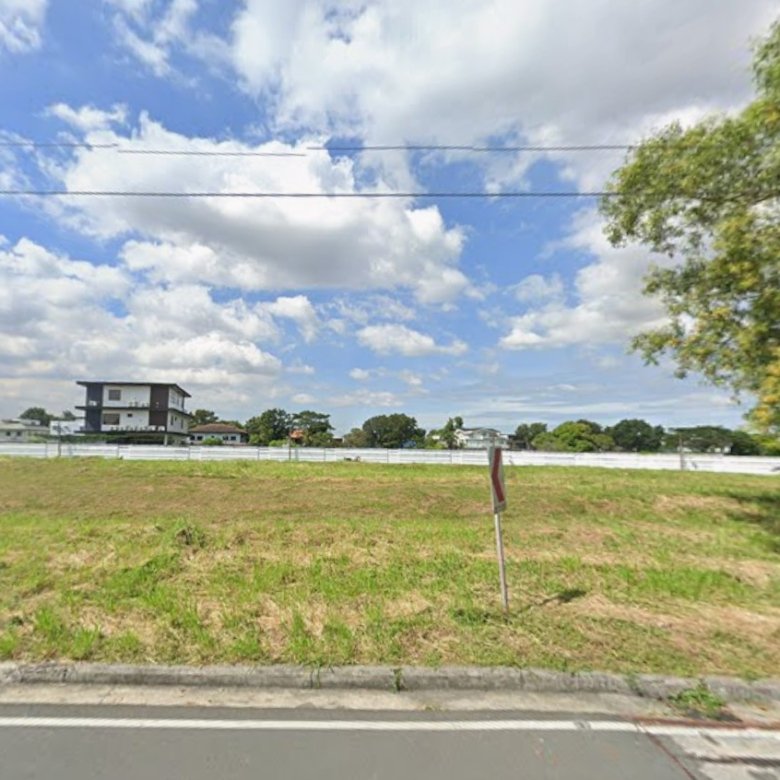 Vacant grassy lot along a street with a white fence, distant buildings, and a large tree on the right under a partly cloudy sky.