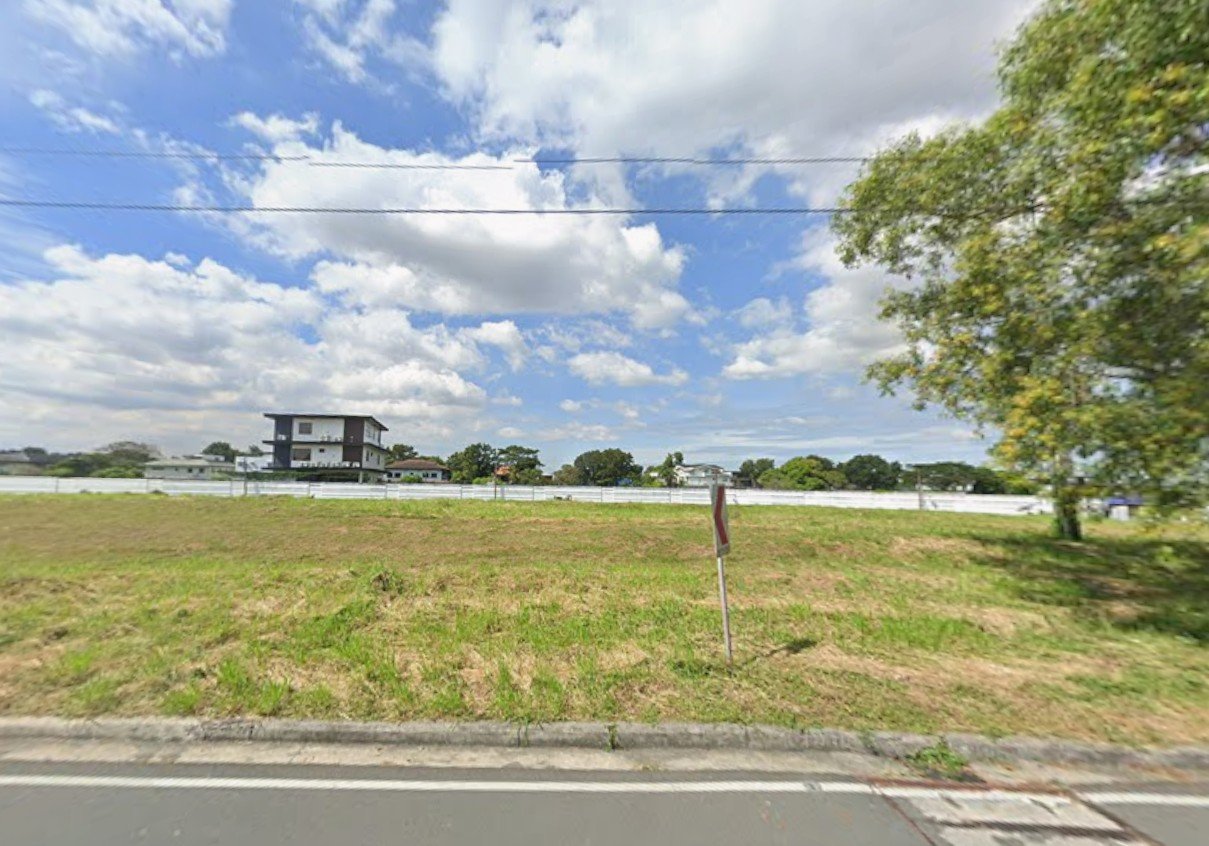 1082 Vacant grassy lot along a street with a white fence, distant buildings, and a large tree on the right under a partly cloudy sky.