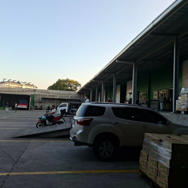 Industrial loading area with stacked pallets, a white SUV in the foreground, and a long green warehouse on the right under a clear sky.