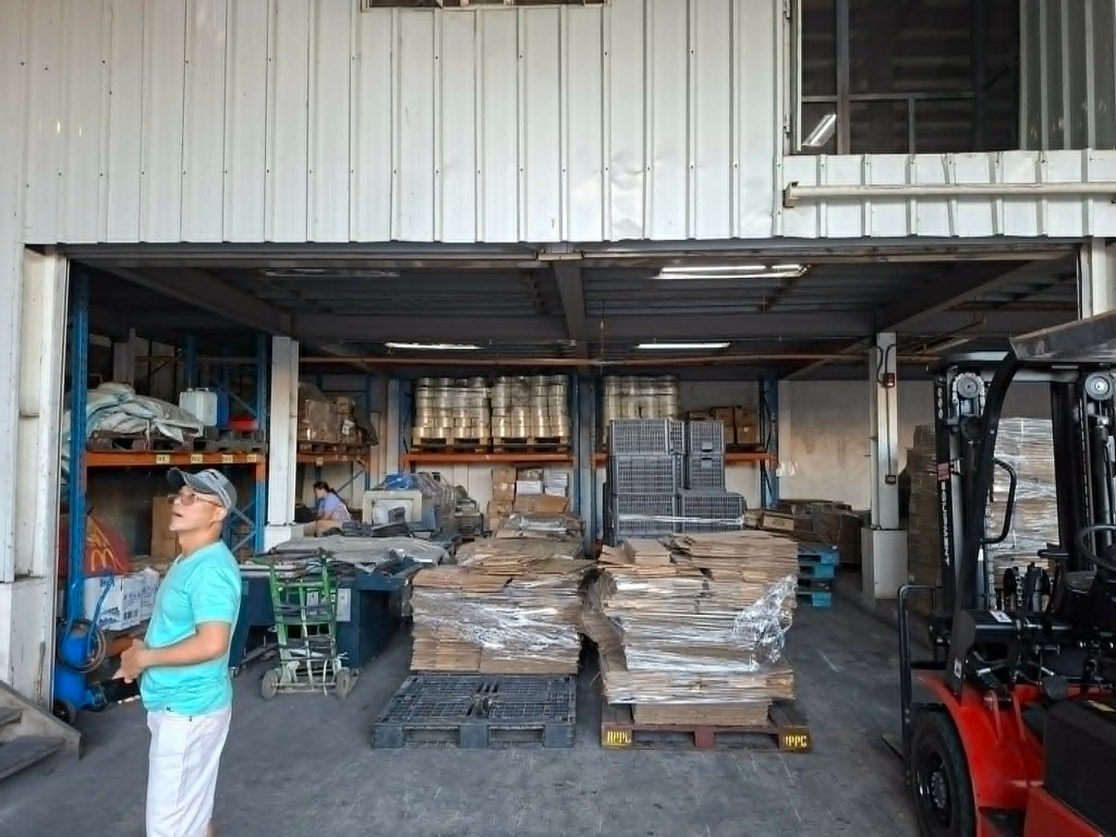 Warehouse with stacked pallets and shelves; a worker in a turquoise shirt stands near the entrance.