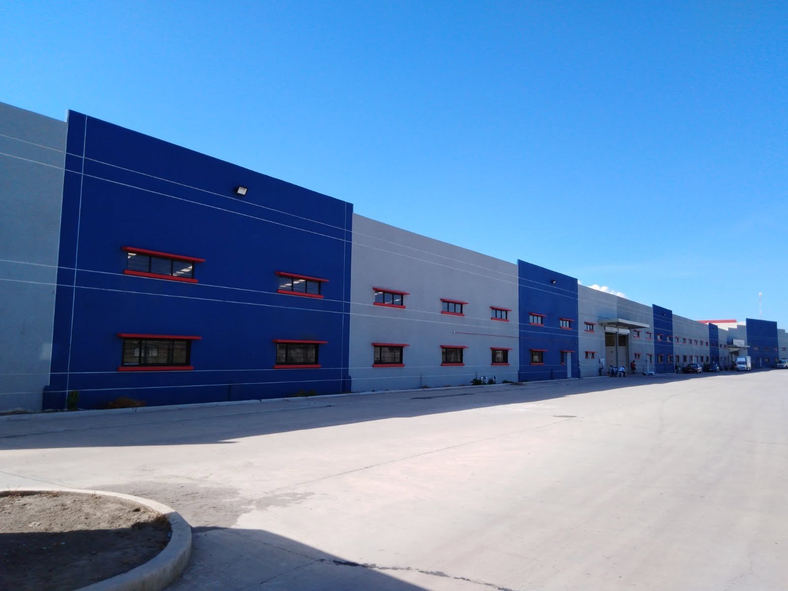 Long industrial warehouse with blue and gray exterior, red-framed windows, under a clear blue sky.