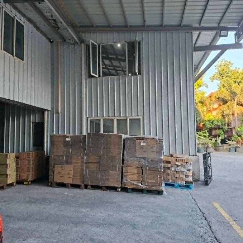 Industrial loading area with stacked cardboard boxes on pallets under a metal canopy; a red car and a white van in the background.