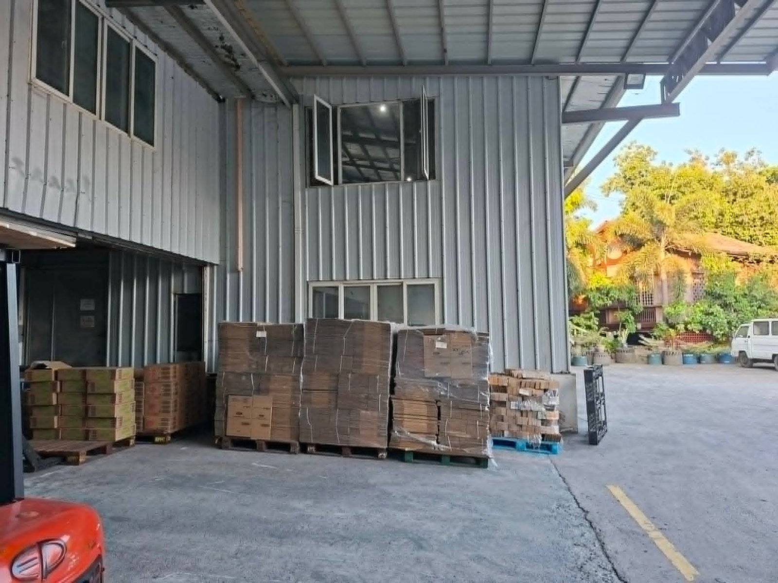 Industrial loading area with stacked cardboard boxes on pallets under a metal canopy; a red car and a white van in the background.