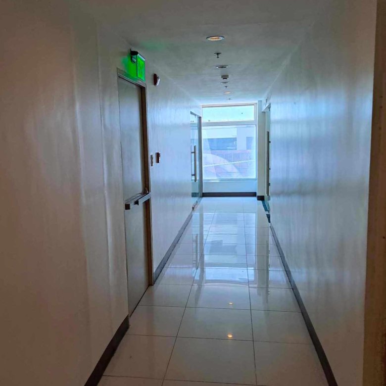 Bright, clean hallway with white tiled floor, doors on both sides, and a green exit sign above a door on the left; natural light at the far end.