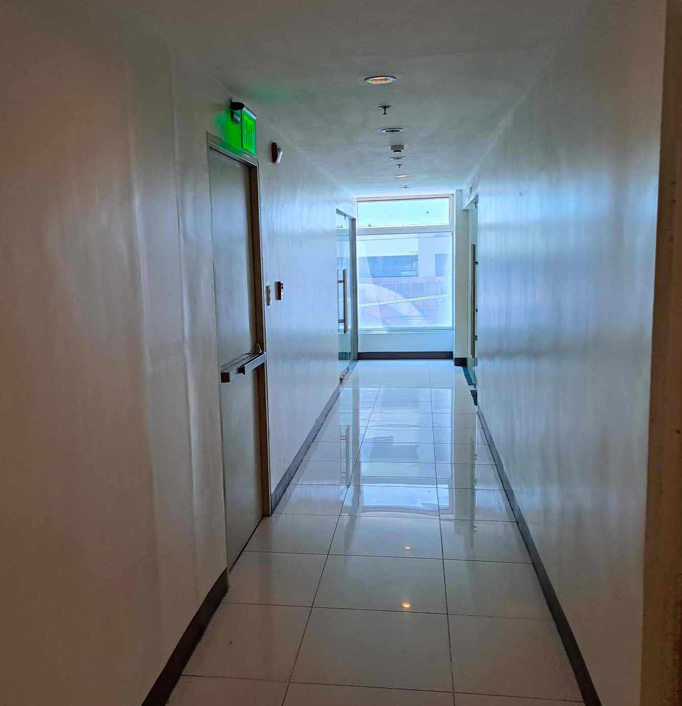 Bright, clean hallway with white tiled floor, doors on both sides, and a green exit sign above a door on the left; natural light at the far end.
