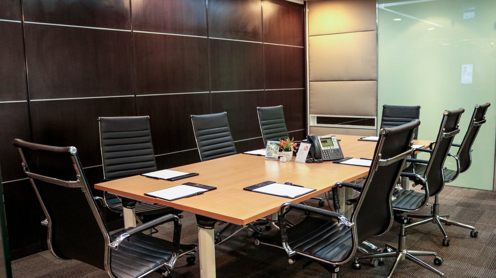 Modern conference room with a long wooden table, black leather chairs, and dark wood wall panels with metal strips on the left.