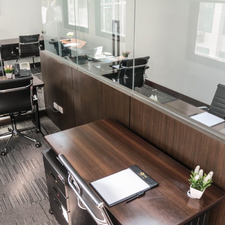 Open-plan office reception featuring dark wood desks, glass partitions, black leather chairs, and small potted plants on the desks.