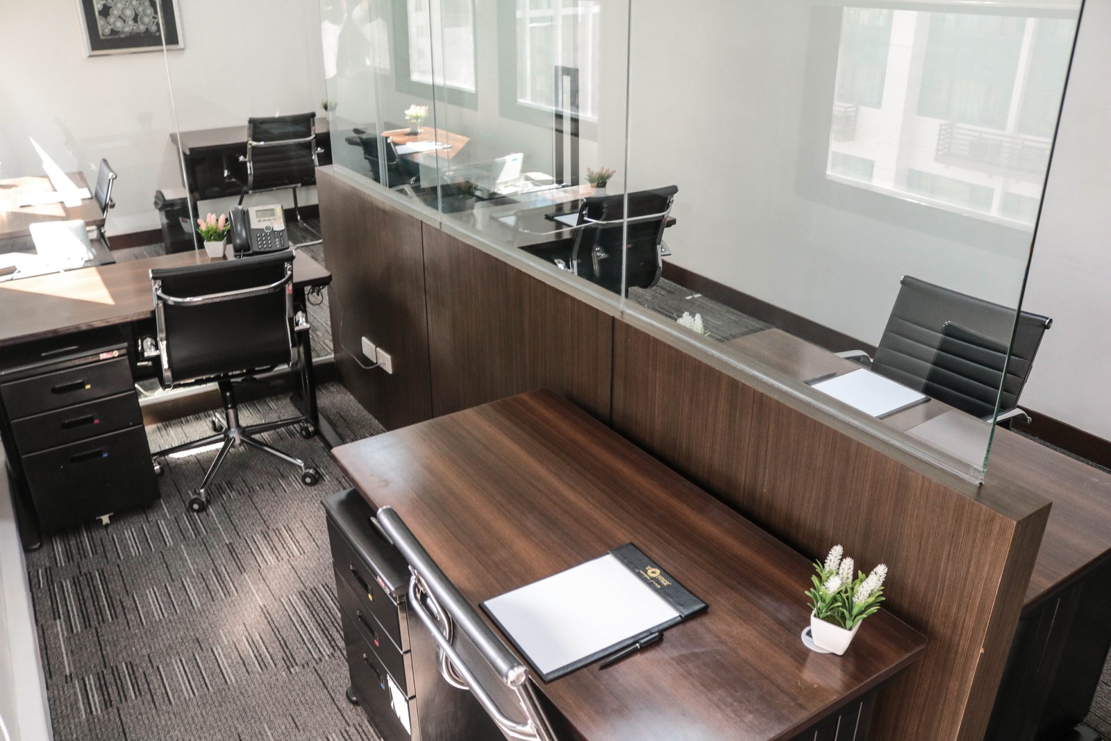 Open-plan office reception featuring dark wood desks, glass partitions, black leather chairs, and small potted plants on the desks.