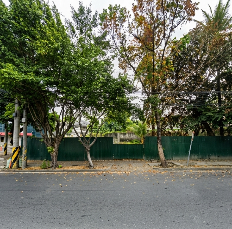 Street view with a row of green trees in front of a green fence and a modern glass building on the left, under an overcast sky, with a paved road in the foreground.