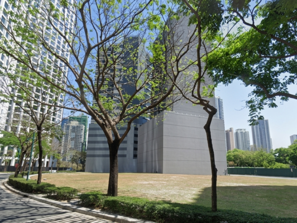 Urban street with a curved road, green trees arching overhead, and tall modern buildings on either side under a blue sky.