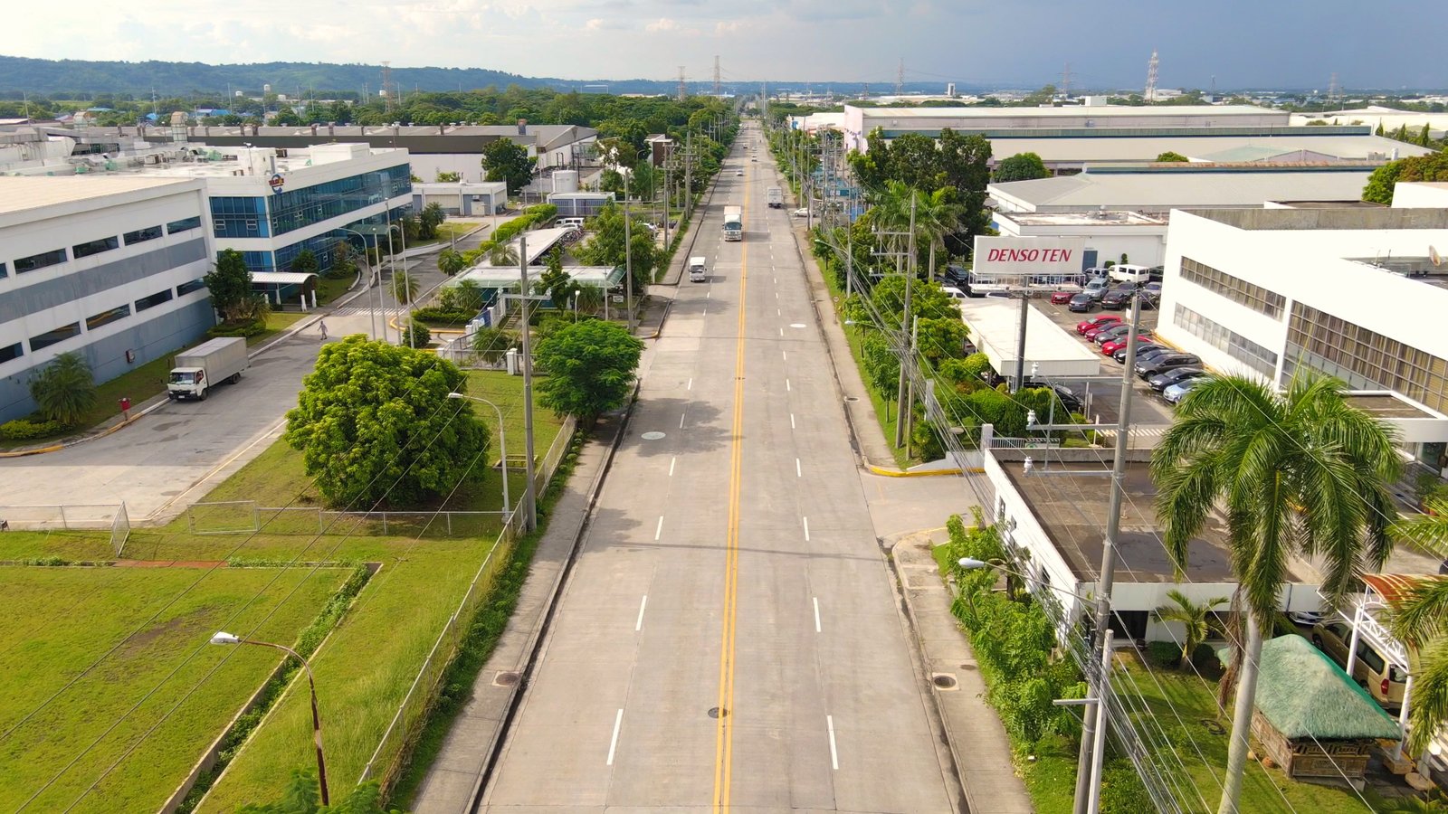 Aerial view of a long street through an industrial area with trees and white buildings, DENSO TEN sign on the right.
