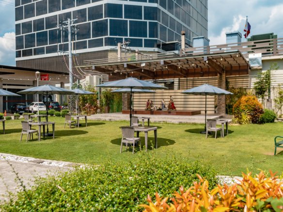Outdoor seating area with tables and blue umbrellas on a green lawn, a modern glass building in the background.