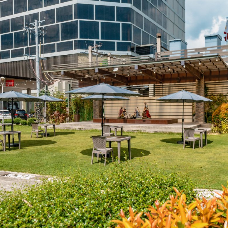 Outdoor seating area with tables and blue umbrellas on a green lawn, a modern glass building in the background.