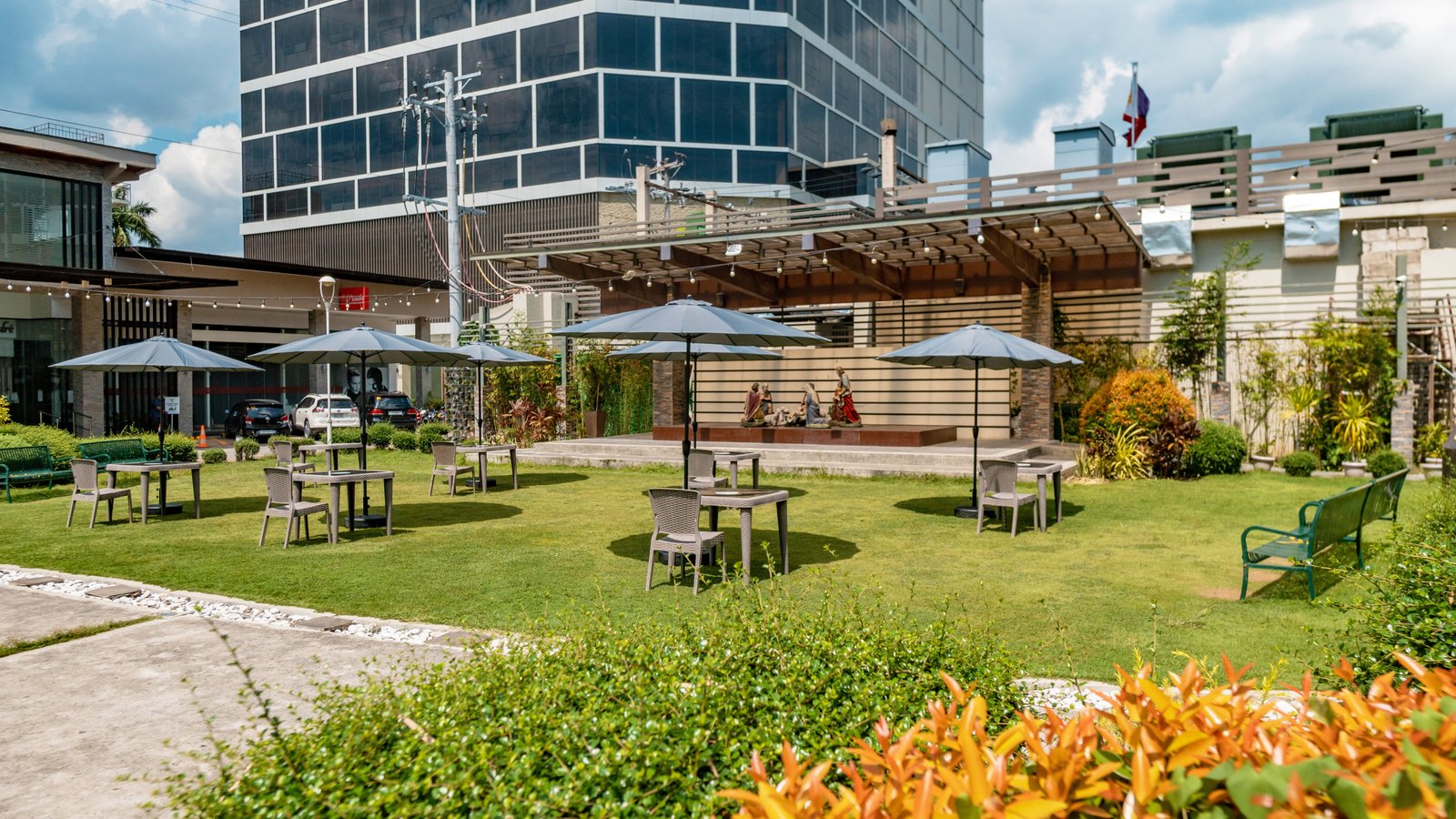 Oakridge Garden Plaza 1 Outdoor seating area with tables and blue umbrellas on a green lawn, a modern glass building in the background.