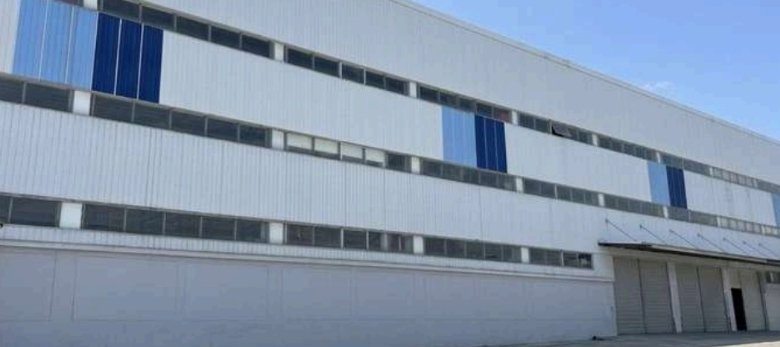 Exterior view of a large industrial warehouse with light gray walls and blue panels, row of loading bays along the bottom, clear blue sky.