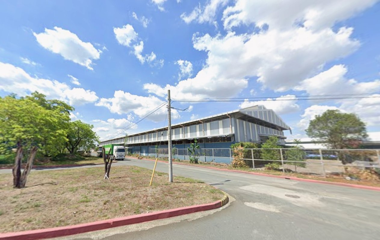 Long modern industrial building along a curved street with a truck at the entrance and a metal fence near the curb.