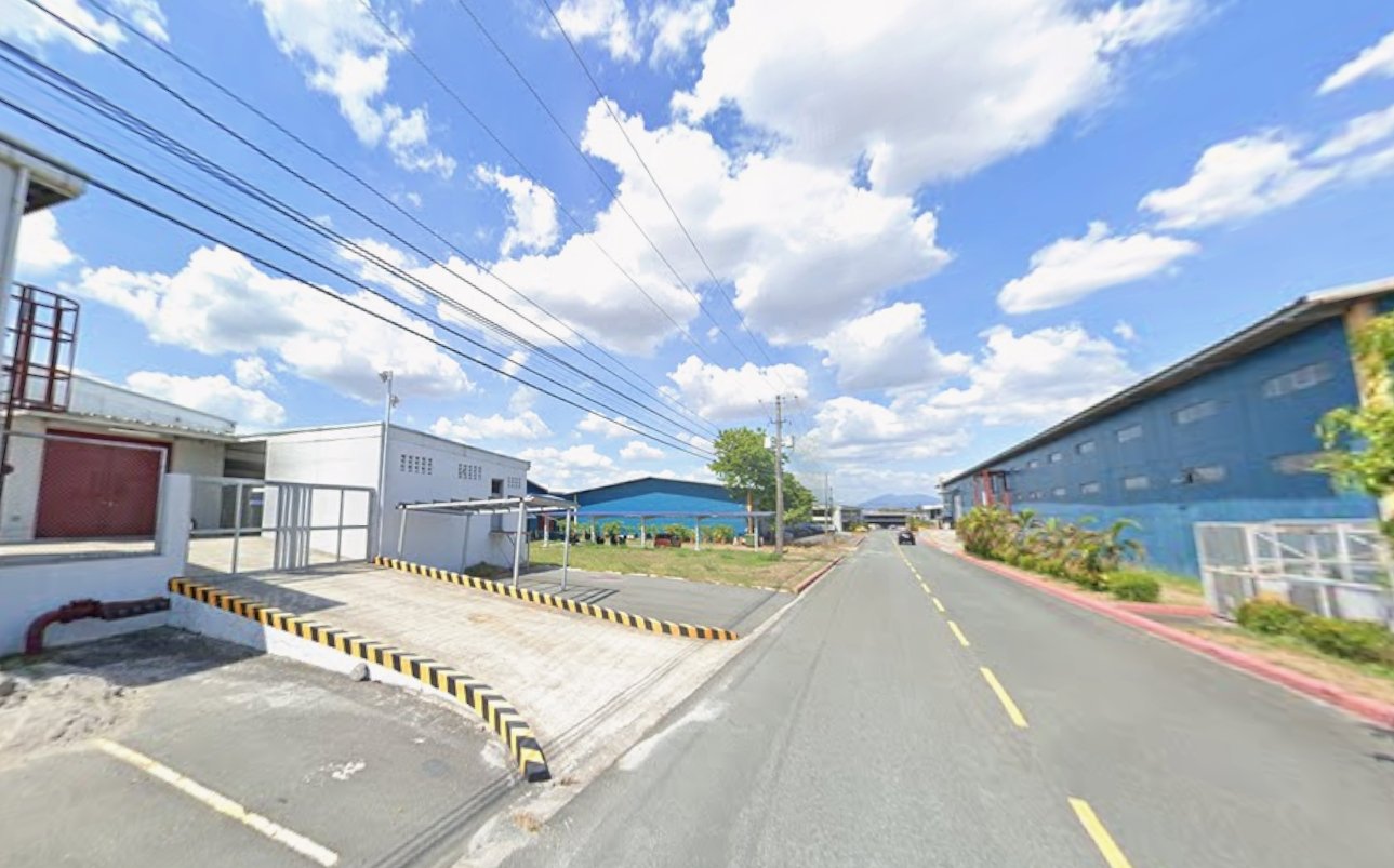 Wide street in an industrial area with blue warehouse buildings, power lines overhead, and a cloudy blue sky.
