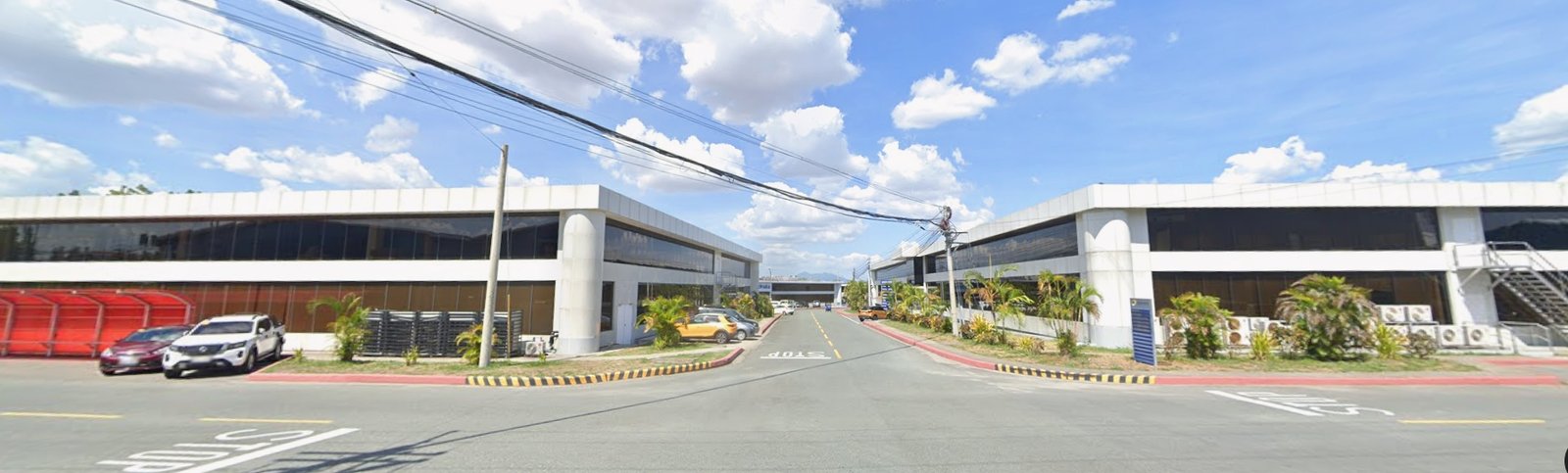 Wide street between two modern white office buildings with glass walls, parked cars along the curb, under a bright blue sky with scattered clouds.