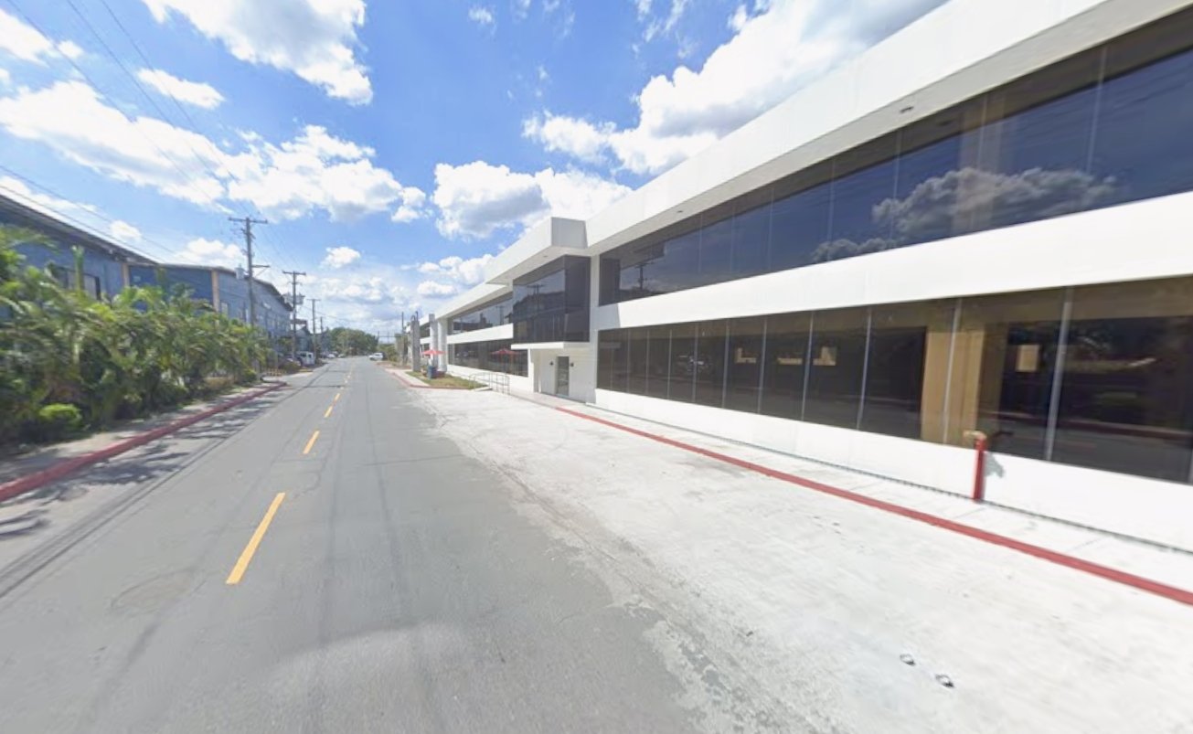 Wide city street with a modern white building and large glass windows on the right, blue sky and scattered clouds above.