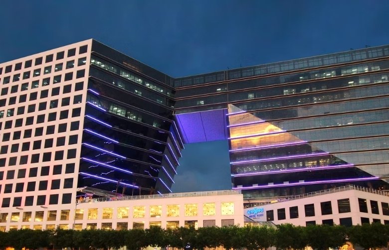 Night view of a two-tower glass building connected by a skybridge, illuminated with purple lights against a dark sky with trees in the foreground.