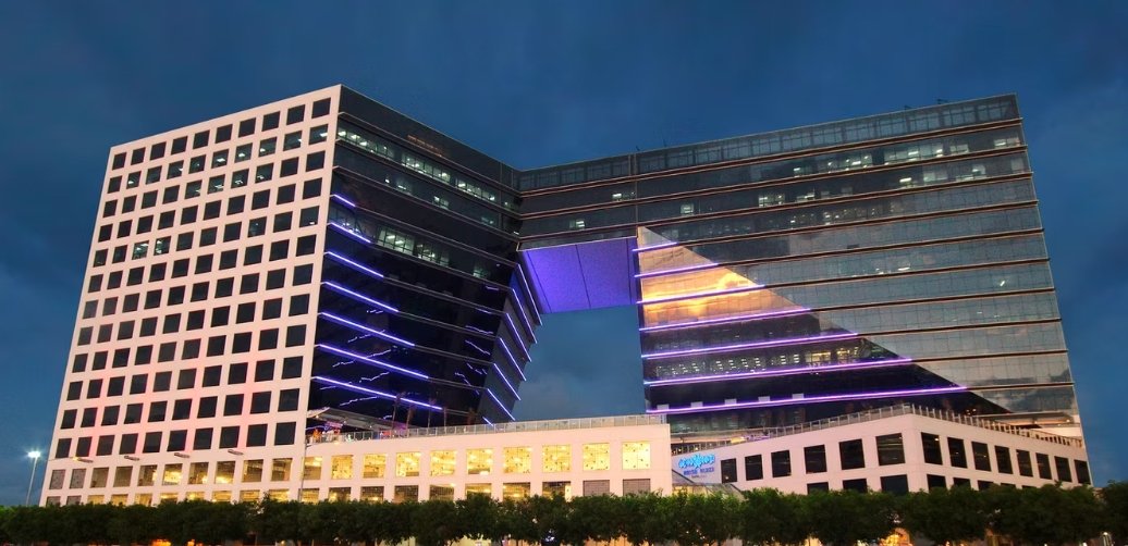 Night view of a modern office complex: two glass towers connected by a skybridge forming a large U-shape, with purple neon lighting and illuminated windows.