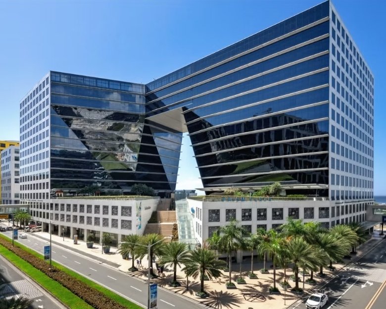 Modern glass office complex with two towers connected by a central archway, palm-lined street in the foreground