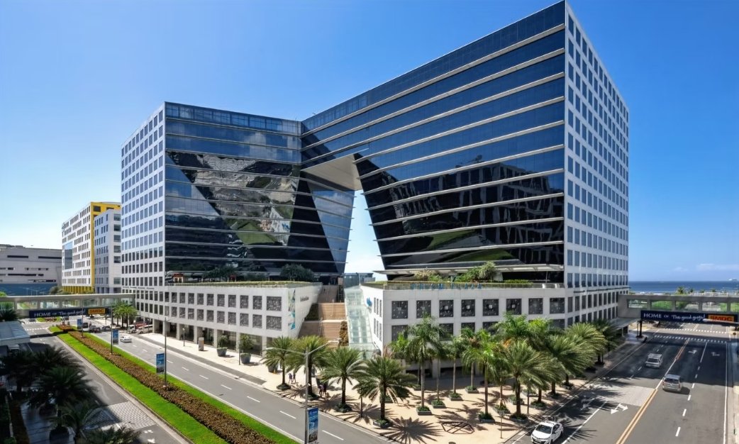 Modern glass office complex with two towers connected by a central archway, palm-lined street in the foreground