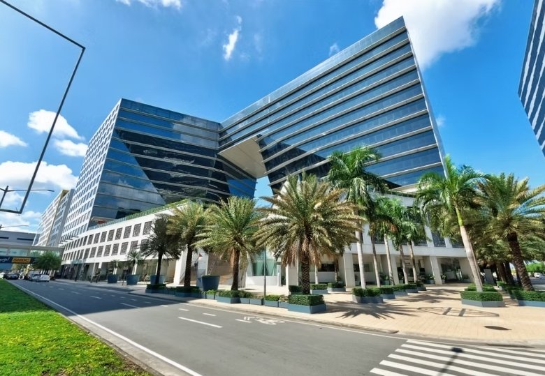 Modern glass office complex with palm trees lining a plaza under a blue sky in daylight to the left of a street edge.