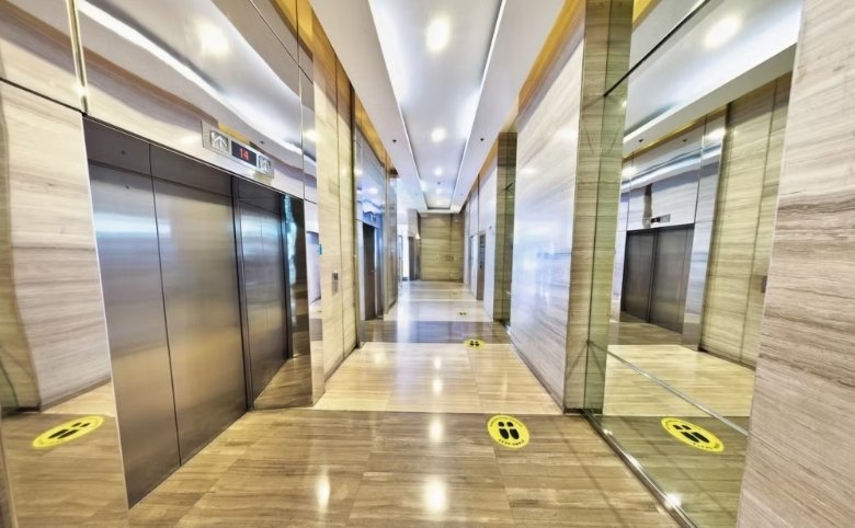 Bright elevator lobby with stainless steel doors, marble walls, and yellow floor markers indicating where to stand for distancing.