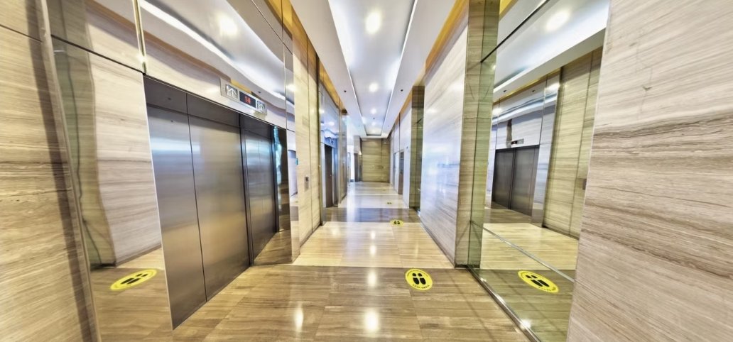 Elevator lobby in a marble-walled hallway with stainless steel doors and yellow floor footprint markers guiding social distancing.”],