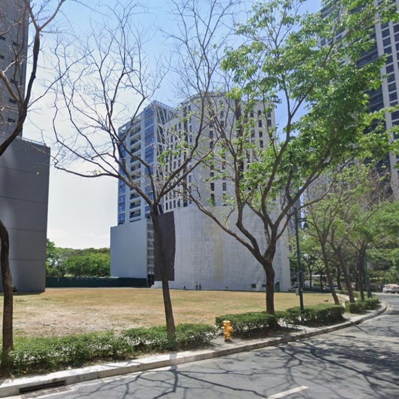 Urban street with a mix of bare and leafy trees, a grassy median, and tall modern buildings under a sunny sky.