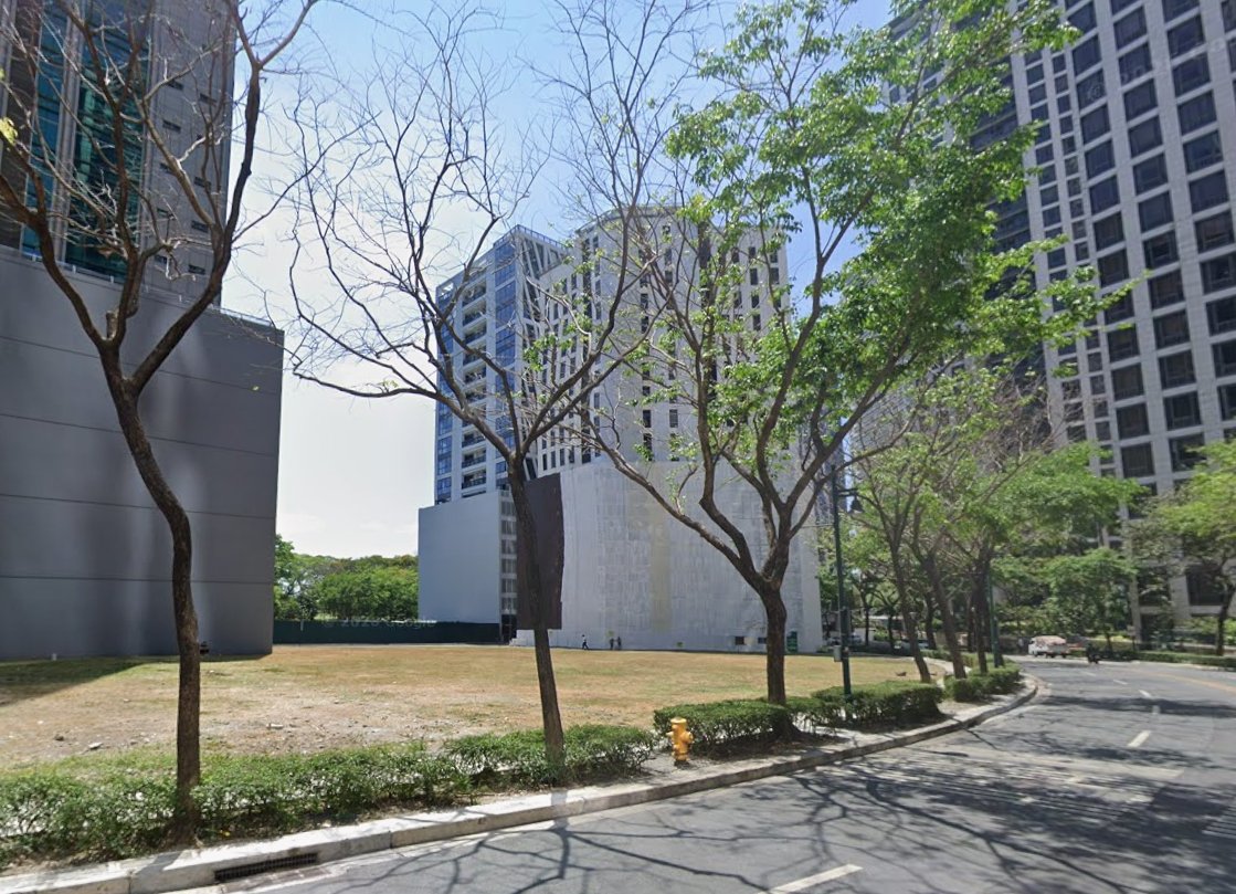 Urban street with a mix of bare and leafy trees, a grassy median, and tall modern buildings under a sunny sky.