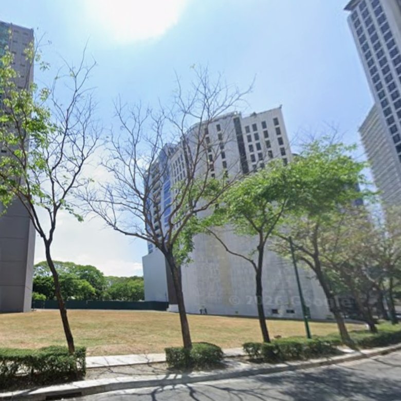 City street scene with tall glass-and-concrete buildings, a row of trees along a curved sidewalk, and a clear blue sky.
