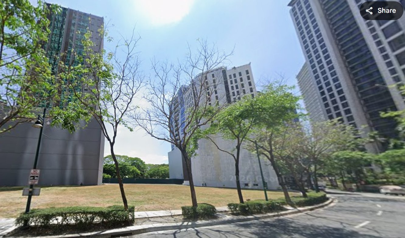 City street scene with tall glass-and-concrete buildings, a row of trees along a curved sidewalk, and a clear blue sky.