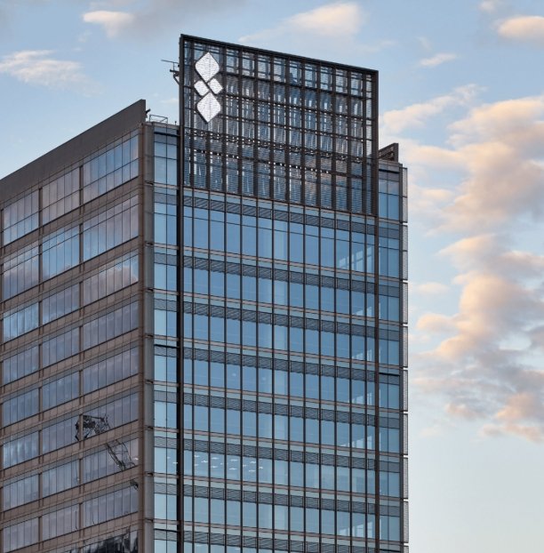 Modern glass office building with a diamond-shaped logo near the top against a blue sky.
