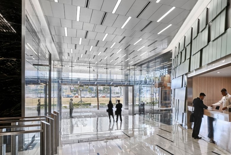 Bright, modern building lobby with a polished floor, glass walls, and a reception desk on the right; people stand near the entrance.