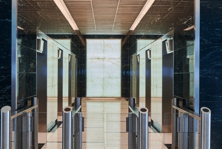 Elevator lobby with mirrored walls, six elevator doors, and stainless steel turnstiles in a marble-lined hall.
